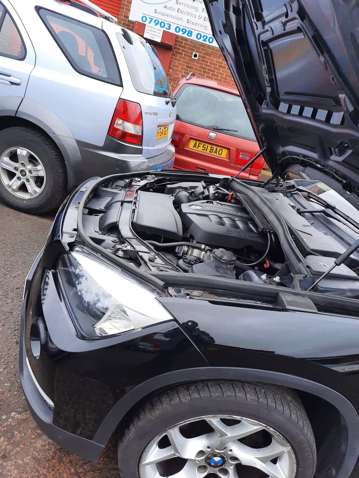 A black BMW with its bonnet open outside the workshop, the Kosi's Car Repairs liveried car parked behind